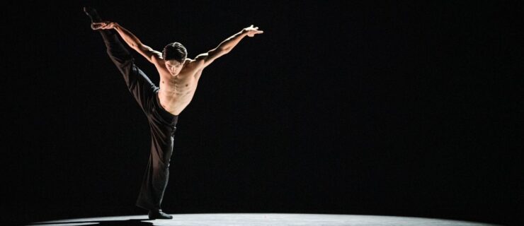 Jun Masuda, lit by a spotlight on a pitch-black stage, lifts one leg a la seconde with a slight bend in his supporting leg. His arms float up in a wide "V" and he looks down to the ground. He is shirtless and wears black pants.