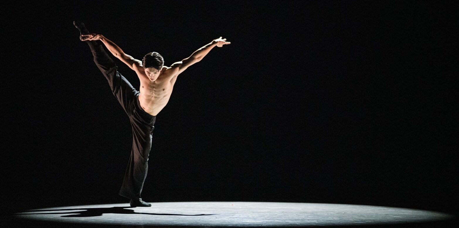 Jun Masuda, lit by a spotlight on a pitch-black stage, lifts one leg a la seconde with a slight bend in his supporting leg. His arms float up in a wide "V" and he looks down to the ground. He is shirtless and wears black pants.