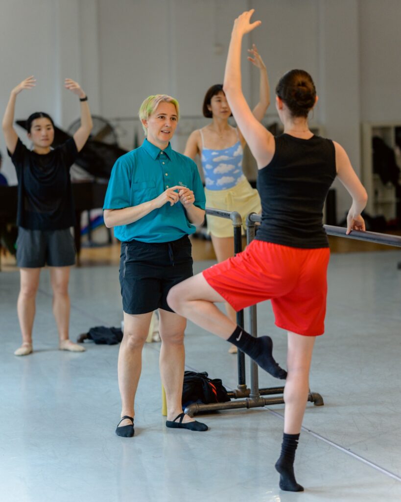 Kade Pyle, in dark shorts, black ballet flats, and a teal button-down, faces a dancer and speaks to them. The dancer, their back to the camera, balances at the barre in retiré en relevé with one arm in high fifth. Two other dancers practice the correction behind Pyle.