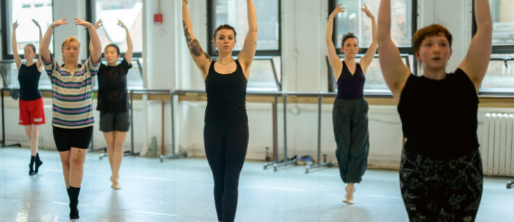 In a brightly lit studio, a class of adult ballet dancers balance in sous-sus with their arms in high fifth. They wear both tight- and loose-fitting practice clothes and flat shoes or socks.