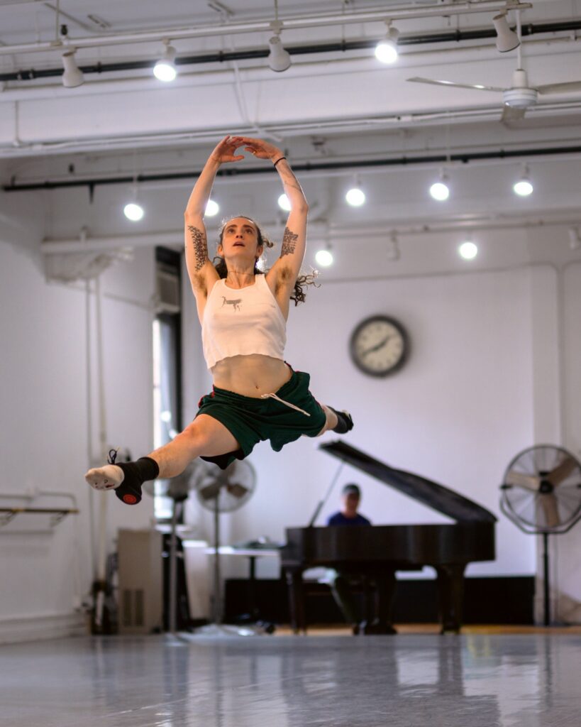 In a ballet studio with portable fans and a grand piano, a dancer does a saut de chat with their arms in high fifth. The dancer wears a cropped white tank top, black athletic shorts, and ballet flats with ankle support.