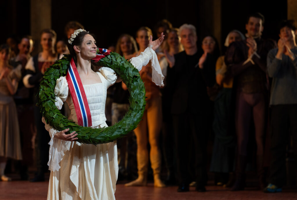 Melissa Hough, wearing a long white dress with puffy sleeves, holds a large wreath around her shoulders and extends one arm out with pride. Behind her, fellow company members clap and celebrate her.