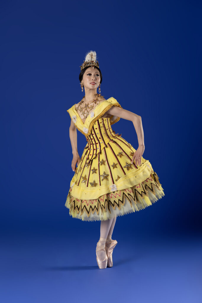 Misa Kuranaga poses on pointe in sous-sus in front of a blue backdrop. She looks out to the side with epaulement and her arms lifted gently to either side, elbows bent, so her fingertips graze her bell-shaped skirt. Her dress is ornate and yellow with gold and dark brown ornamentation. She wears a gold necklace with stars and a feathered headpiece.