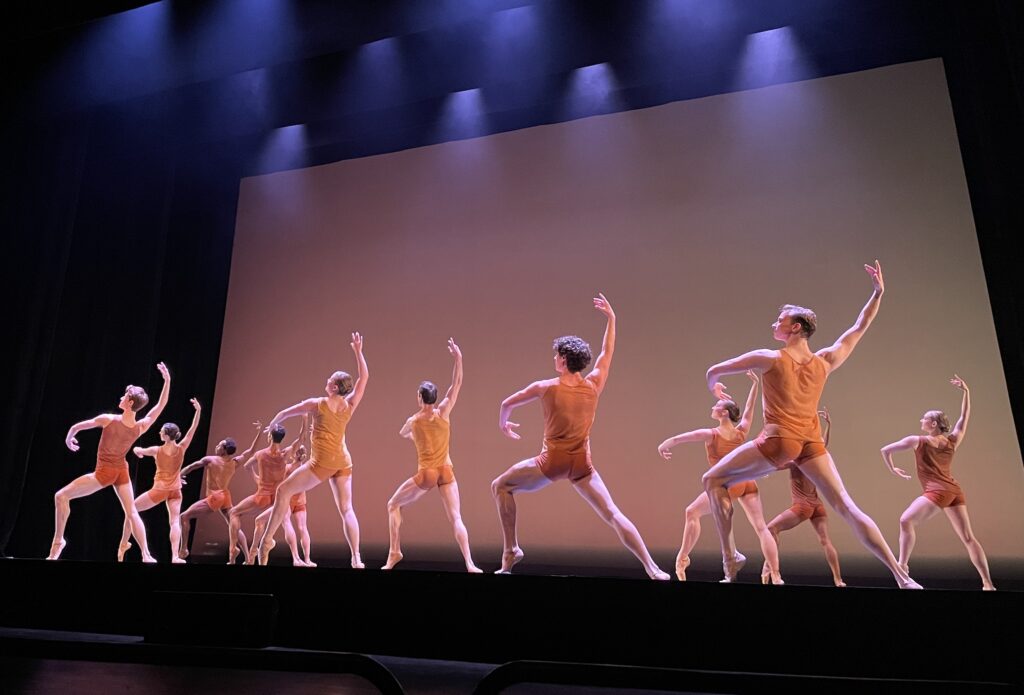 A large corps of male and female dancers pose in profile facing stage right. They stand in a wide lunge with their left foot in front, popped up onto demi-pointe or pointe. They twist their upper bodies with their left arms curved in front of them and their right arms lifted. All wear orange tank tops and matching shorts.