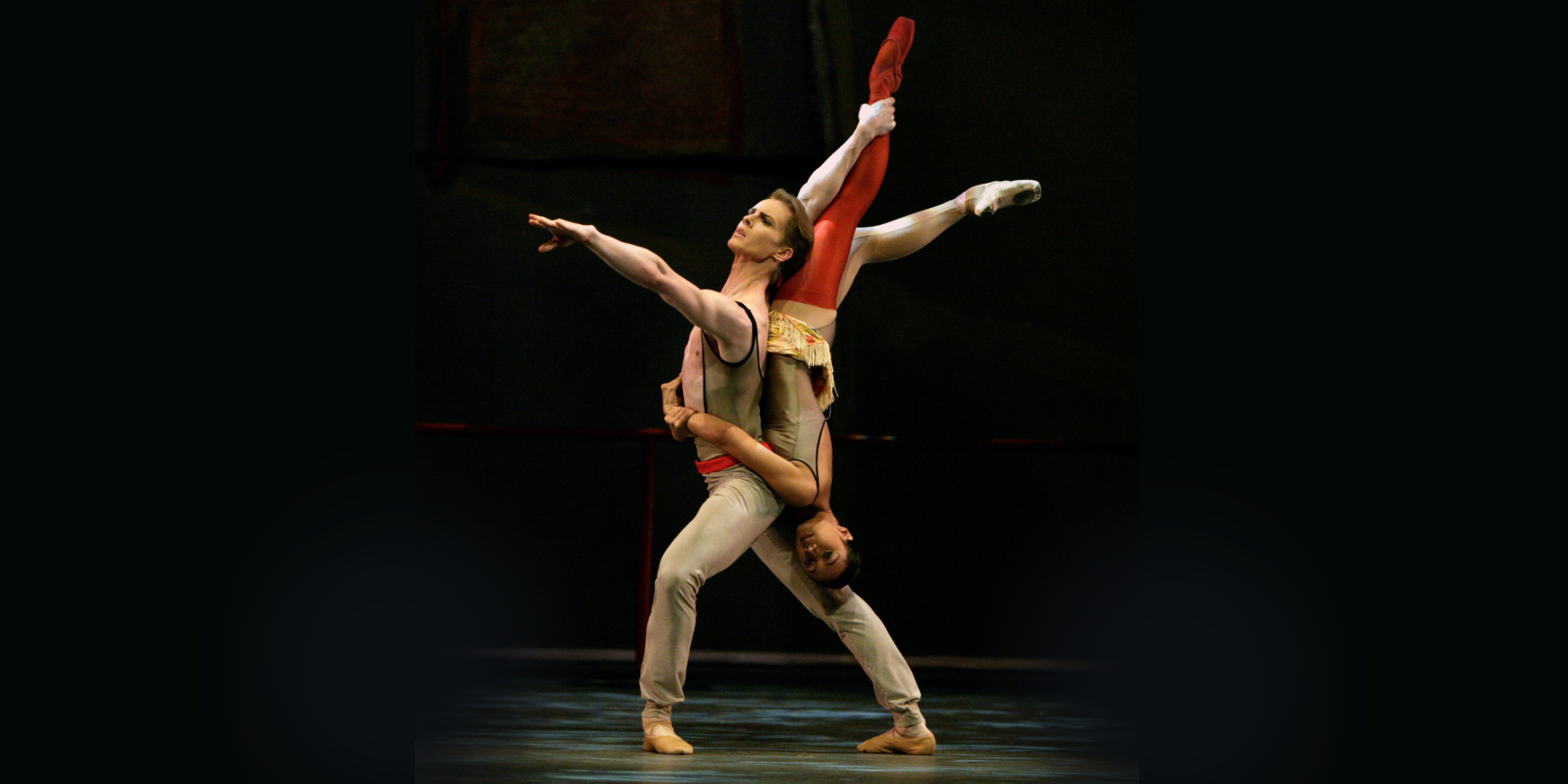 Onstage, Tiit Helimets lunges forward while holding Ommi P. Leffler upside-down, using one arm to hold one of her ankles as his other extends forward. Leffler wraps both arms around Helimet's waist, her head by his back thigh, as she extends one leg vertical and one behind, with a slight bend.