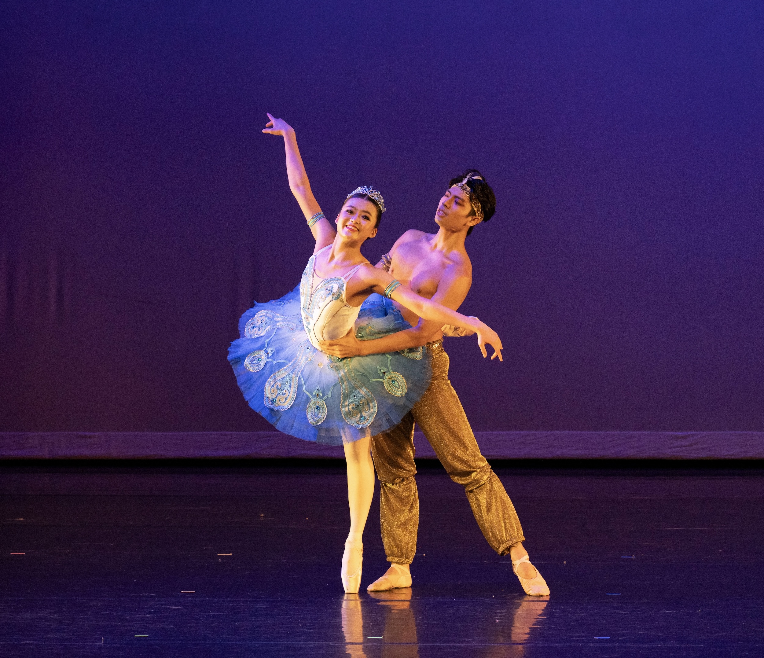 Jessica Wang poses in arabesque on pointe facing the audience as Song Teng partners her around the waist. Wang wears a blue and white tutu with one of her blue tiaras, and Song wears flowy tan pants.
