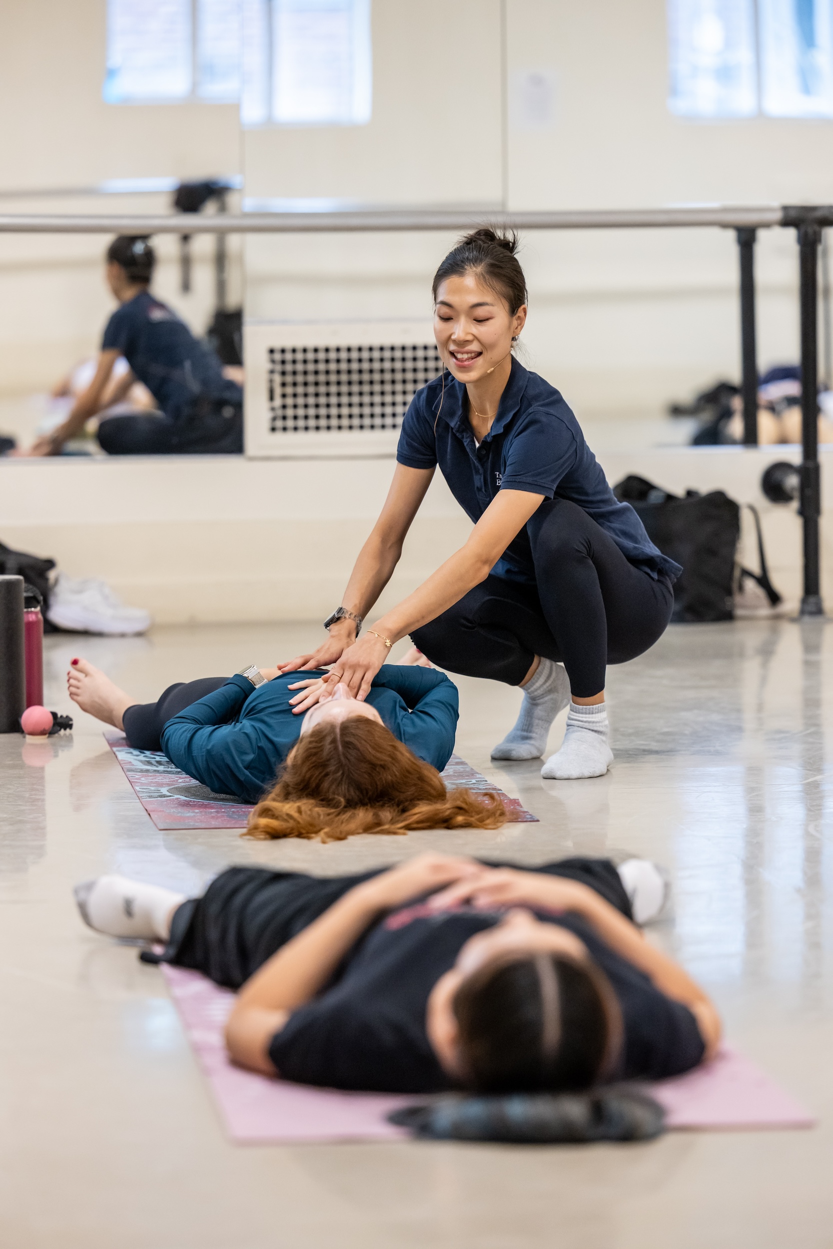 SeHyun Jin, wearing a blue polo shirt and black leggings, adjusts a dancer lying on a yoga mat. The dancer holds their right hand to their chest and their left hand on their belly.