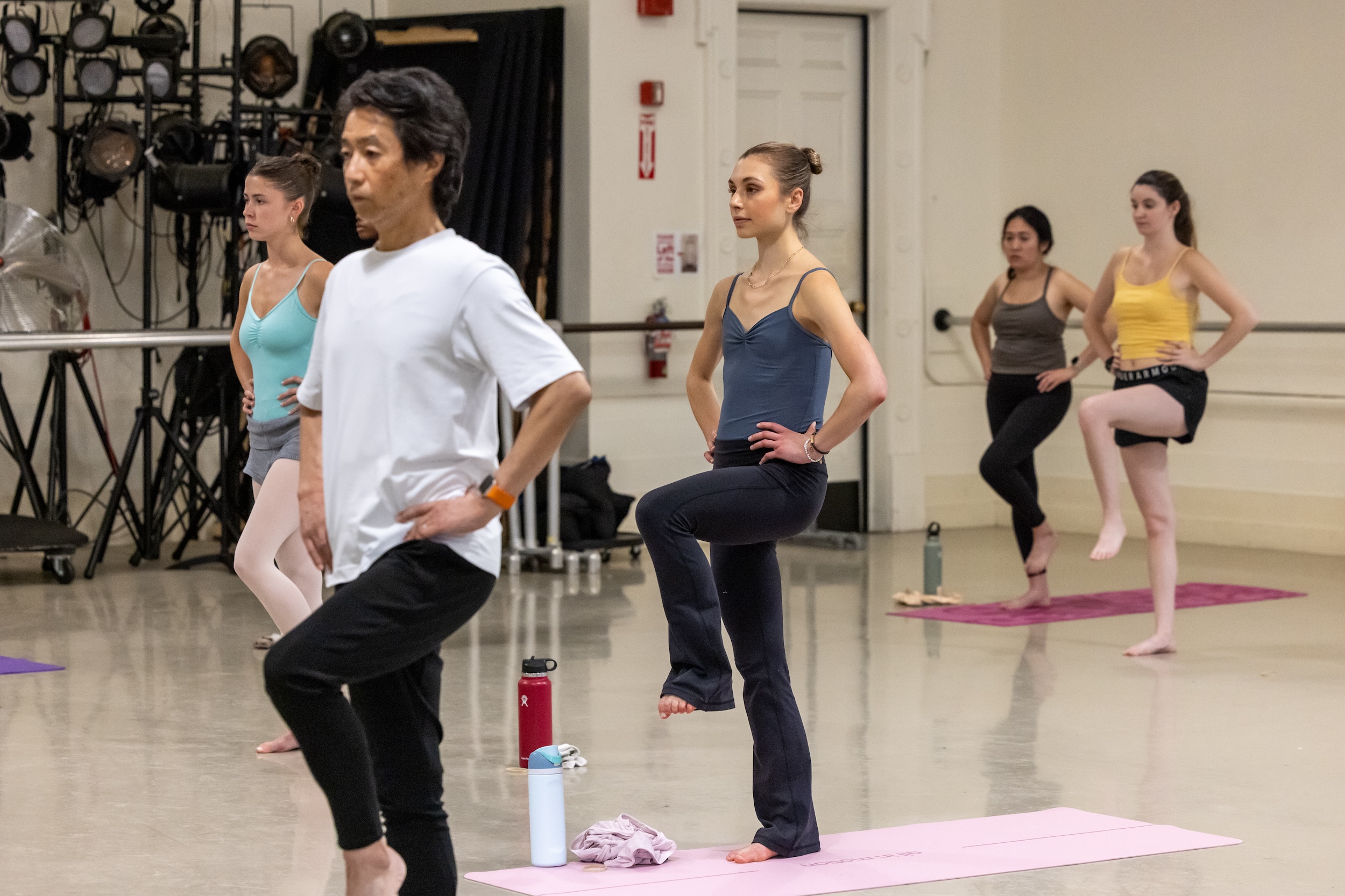 A group of adults wearing dancewear stand on yoga mats in a large dance studio. They lift their left foot off the ground, balancing with their hands on their hips.