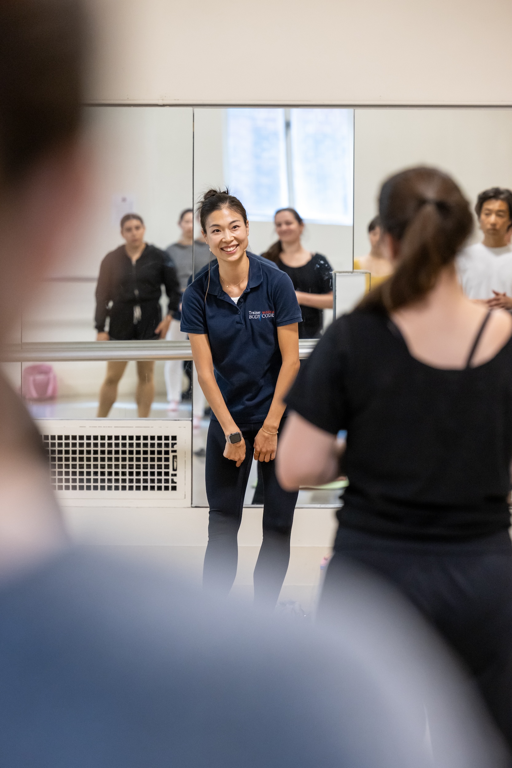 SeHyun Jin stands in the center of a ballet studio and smiles as she addresses a class of students. She wears a blue polo shirt and black leggings.