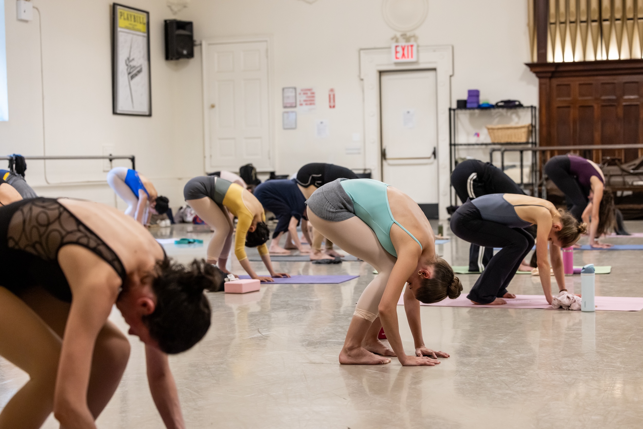 A group of dancers stand on yoga mats and bend down, touching the floor with their hands as they bend their knees.