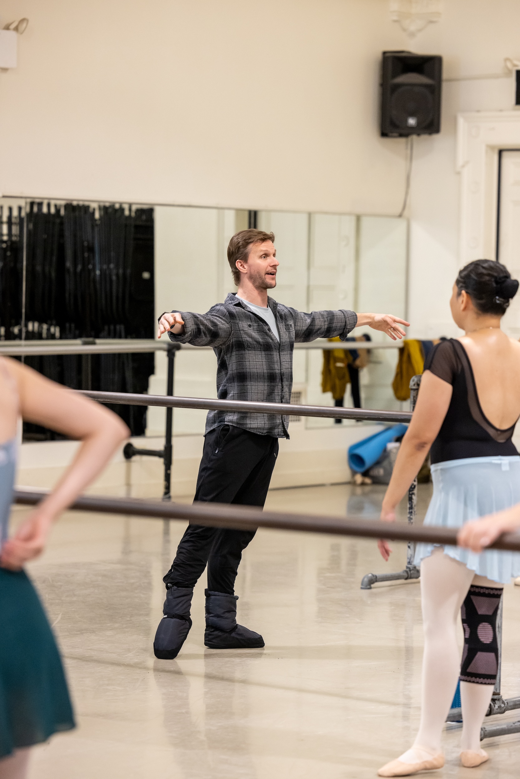 Daniel Ulbricht, wearing a flannel shirt, black pants, and warm-up booties, demonstrates a tendu à la seconde at the barre during a ballet class.