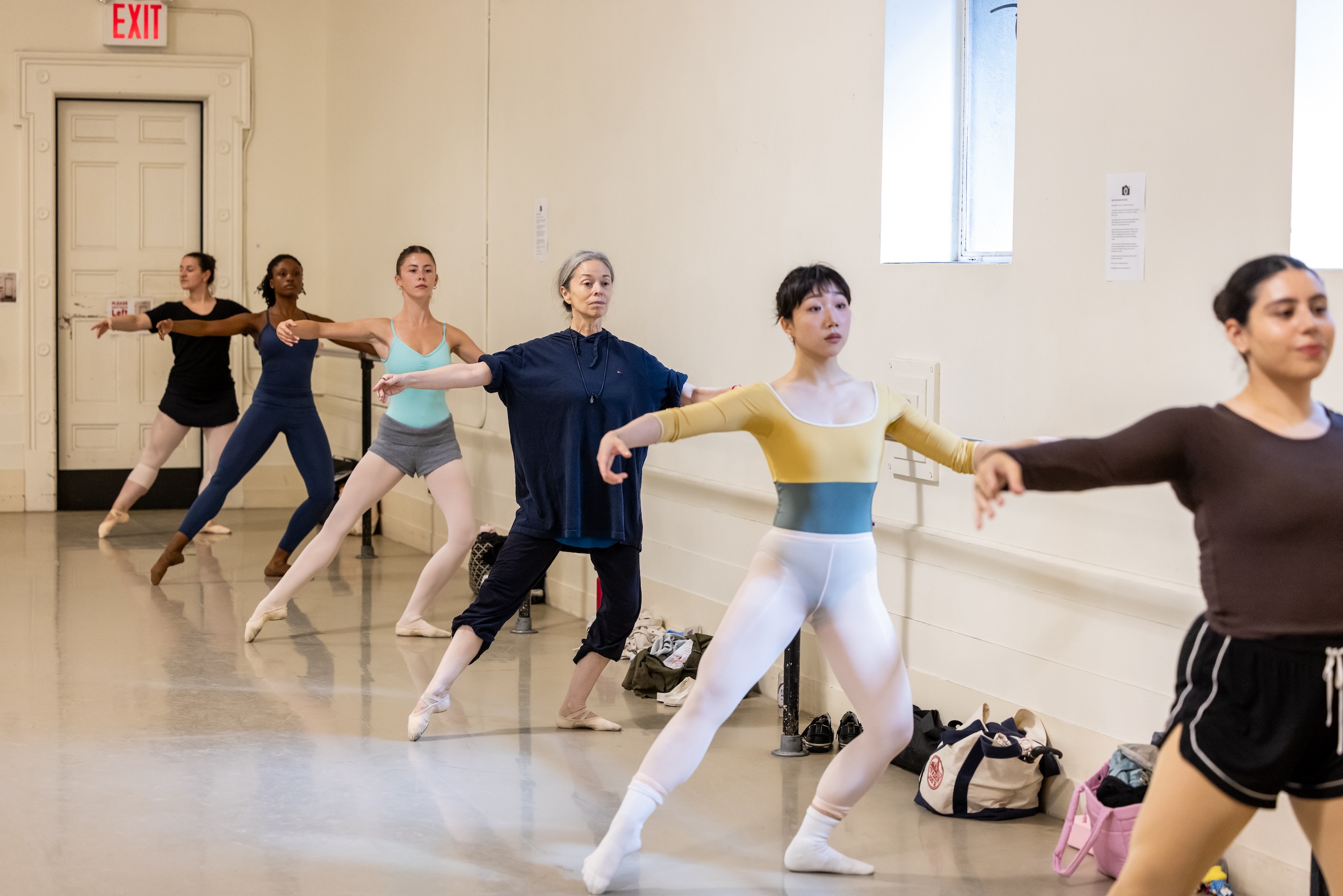Six adult ballet dancers wearing various practice clothes to a tendu à la seconde at the barre, with their supporting leg in plié.