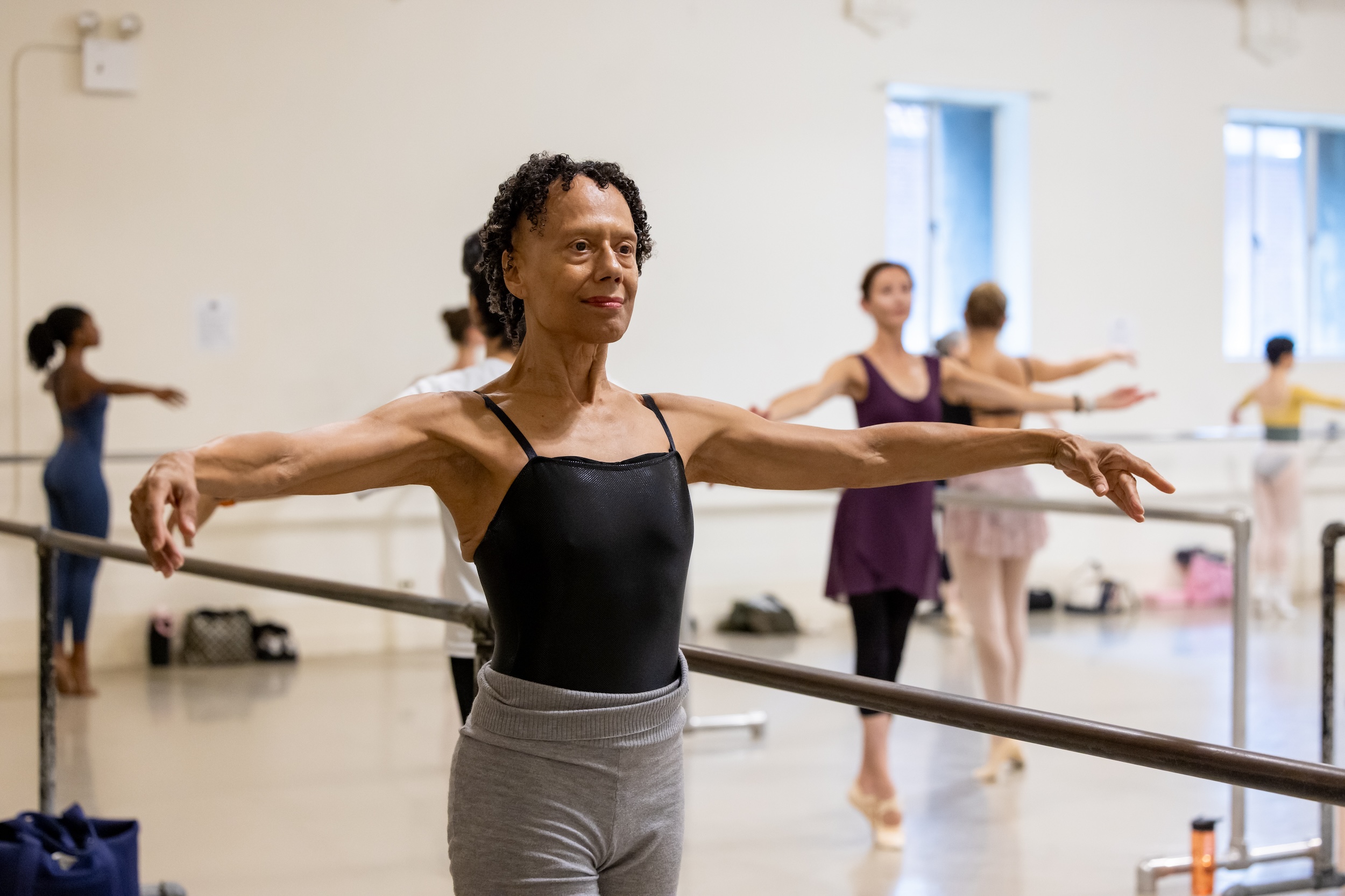 A woman wearing. ablack camisole letard and gray warm-up pants is shown at the barre from the thighs up with her arms in second position. Other dancers are in the background balancing in sus-sous, also with their arms in second position.