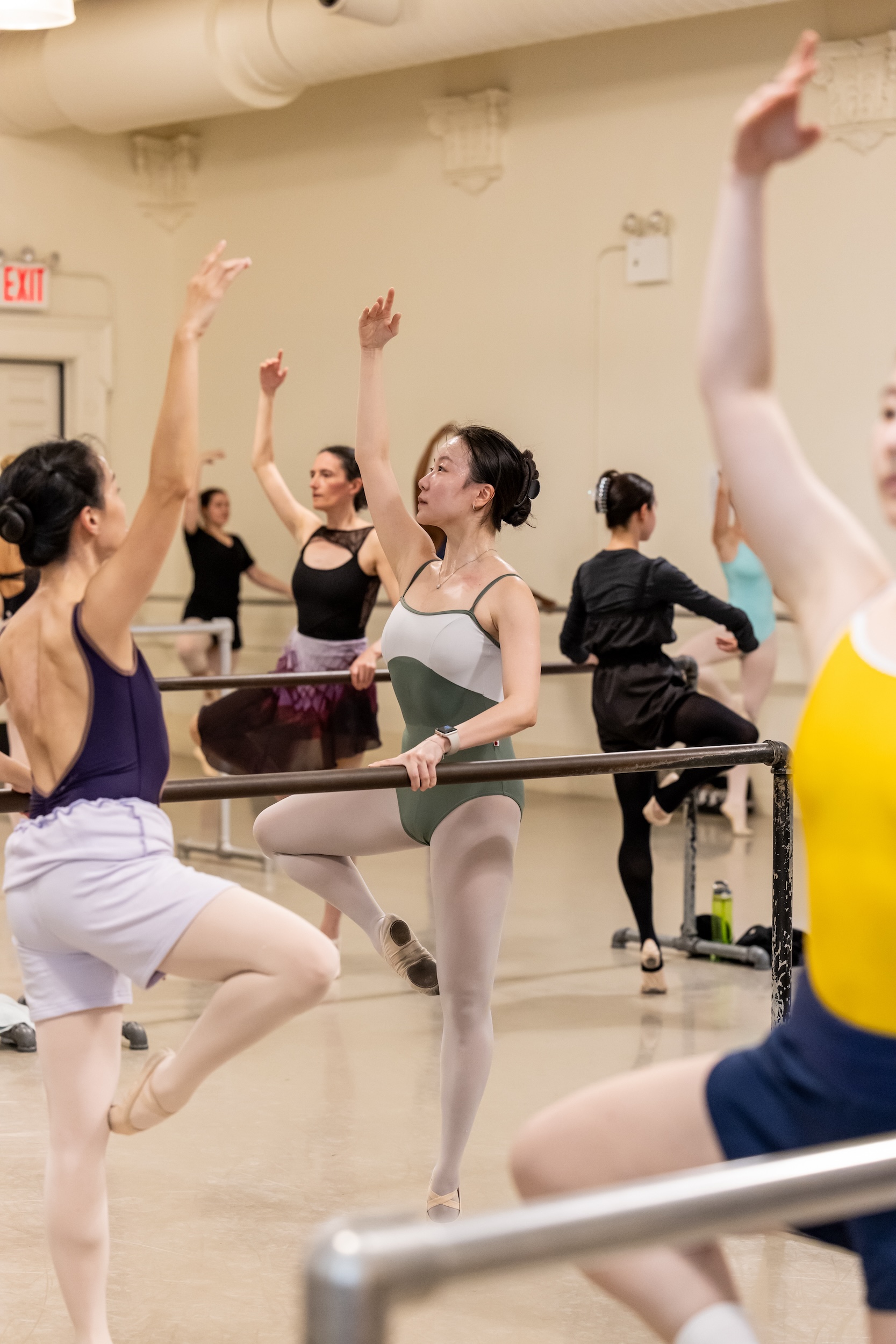 A class of adult ballet dancers wearing various leotards, tights, warm-ups, and ballet slippers, practice retiré at the barre.