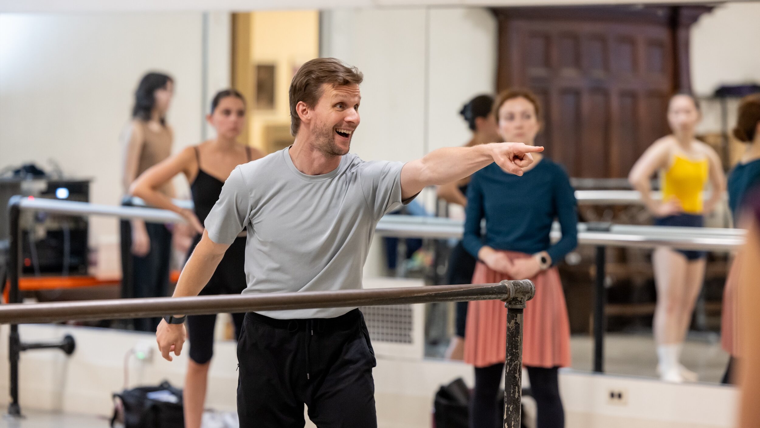 Damiel Ulbricht, wearing a gray t-shirt and black pants, smiels and points to someone out of view during a ballet class. Students in various practice clothing stands in the background, watching.