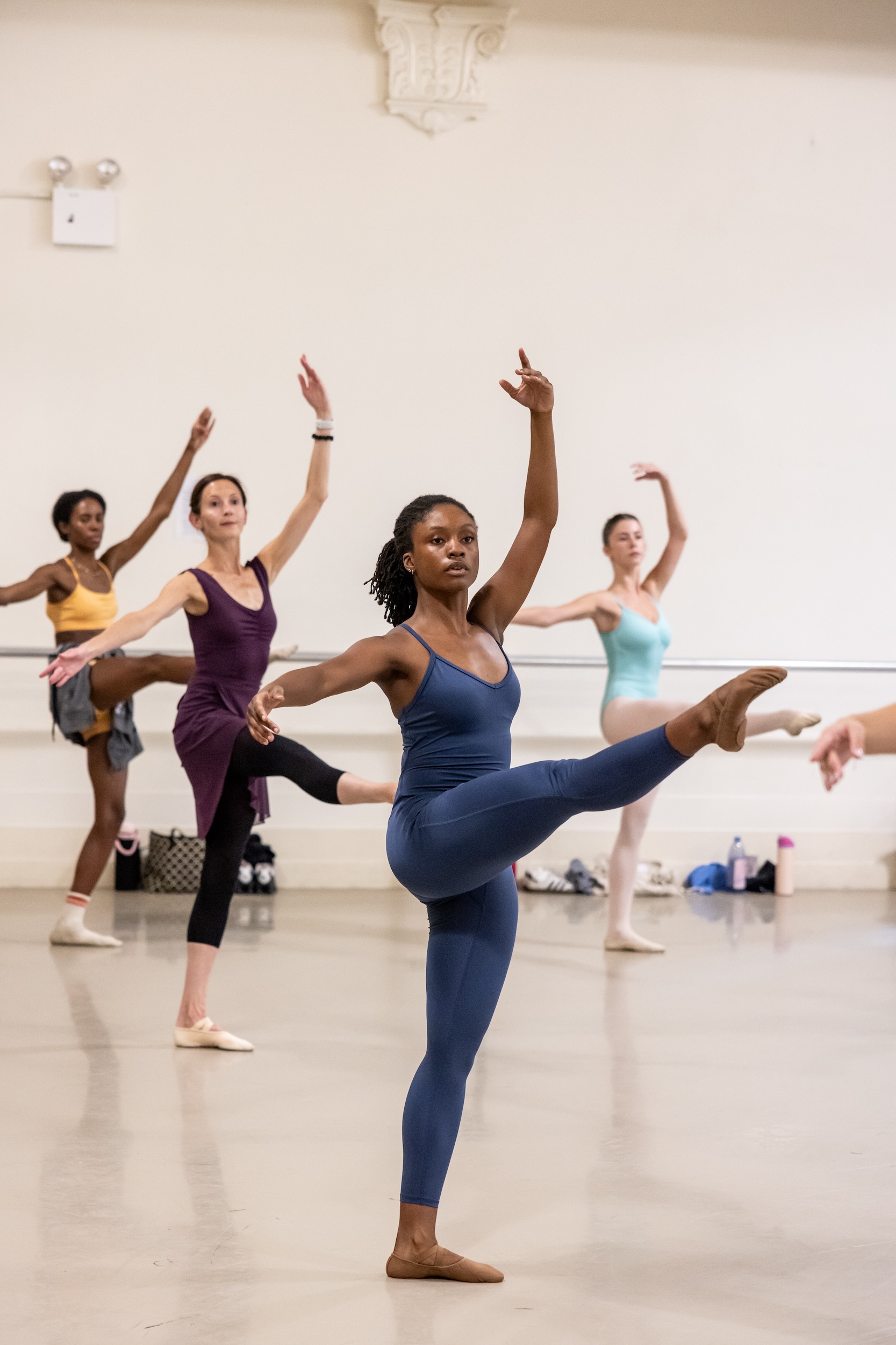 An adult woman in a blue unitard and brown ballet slippers practices a développé in croisé devant during an adagio exercise. Several other dancers practice the same step behind her.