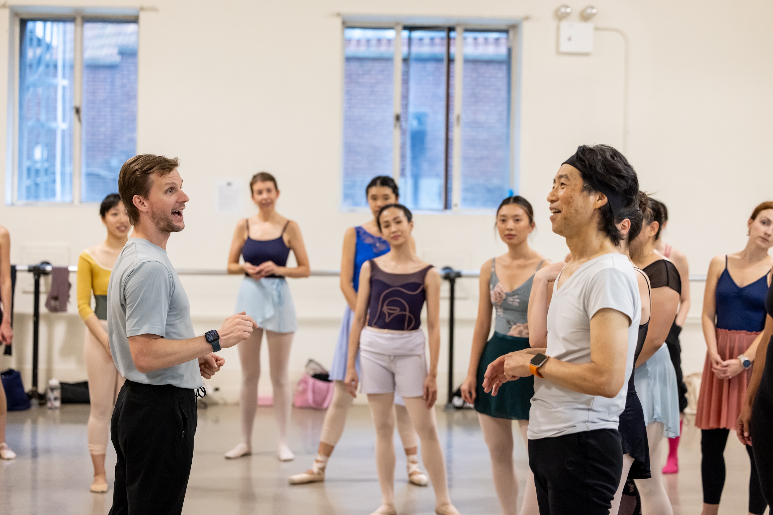 Daniel Ulbricht, wearing a light-colored tishrt and black pants, talks and laughs with a group of adult ballet students in a large dance studio.