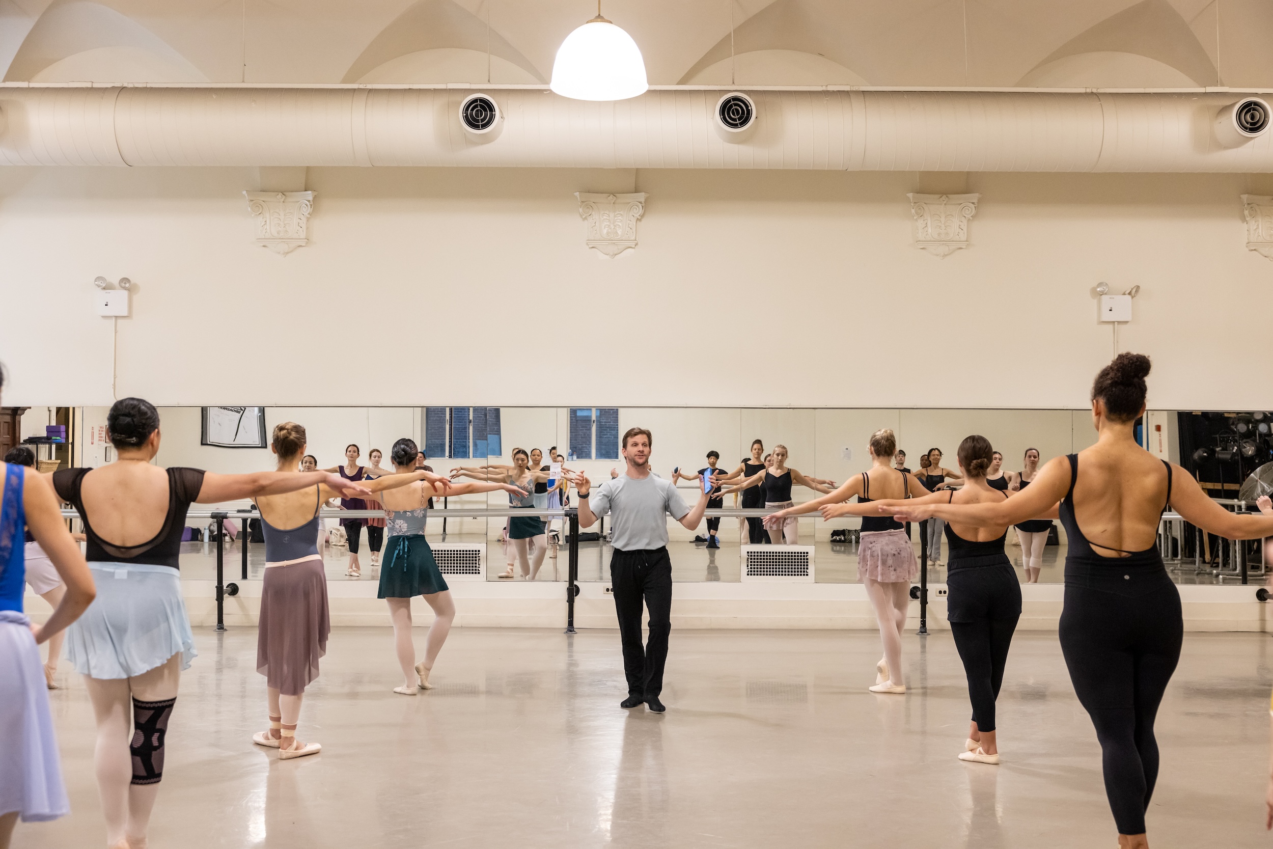 Daniel Ulbricht stands in the middle of the dance studio, facing a large group of dancers wearing practice clothes. He holds his hands up as if to cue music as the dancers open their arms out to the side.