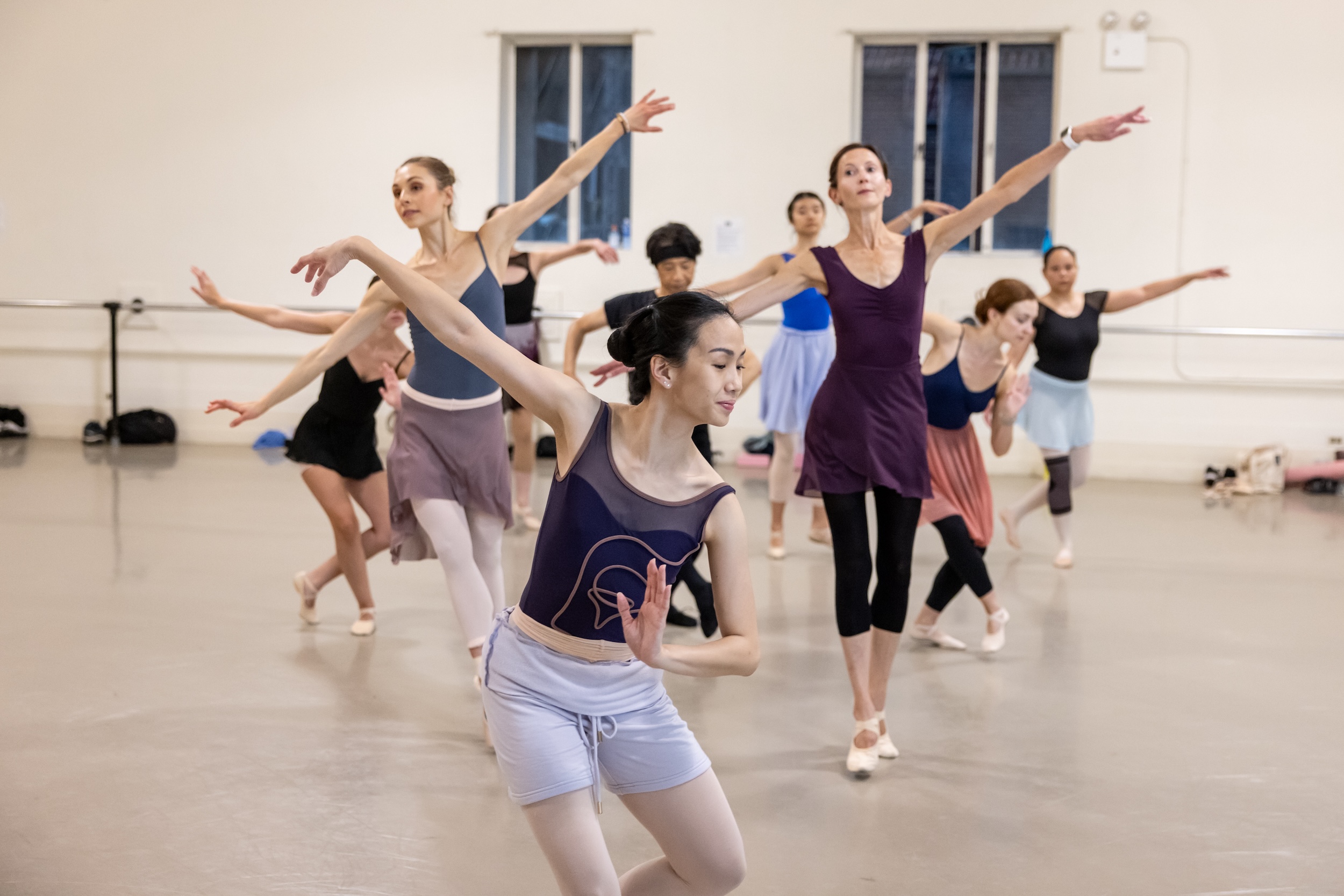 A group of adults dancers wearing rehearsal clothing practice choreography in a large dance studio.