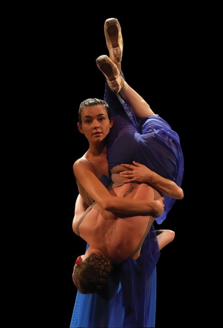 Rachele Perla, shown from the knees up, faces front while holding an upside-down Amber Neff around her middle. Neff leans slightly so her head rests on Perla's opposite hip, wrapping her arms around Perla's waist as she bends her legs slightly. They wear long blue dresses.