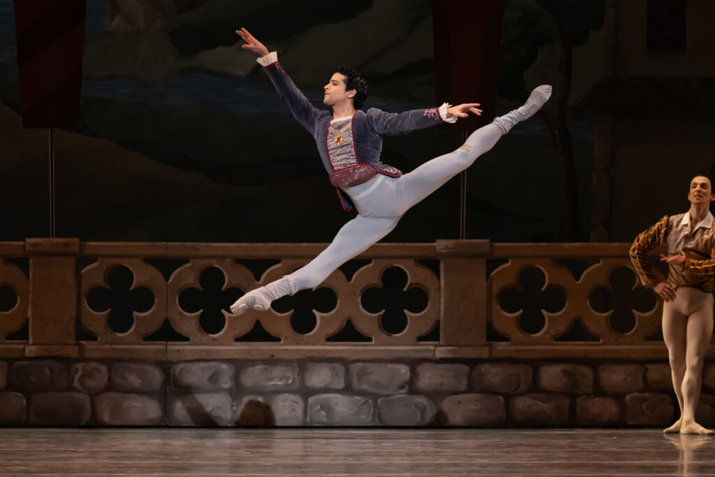 Rafael Quenedit splits his legs in the air several feet above the ground. He reaches his arms in a high arabesque, looking forward toward stage left. He wears light gray tights with a dark blue velvet vest.