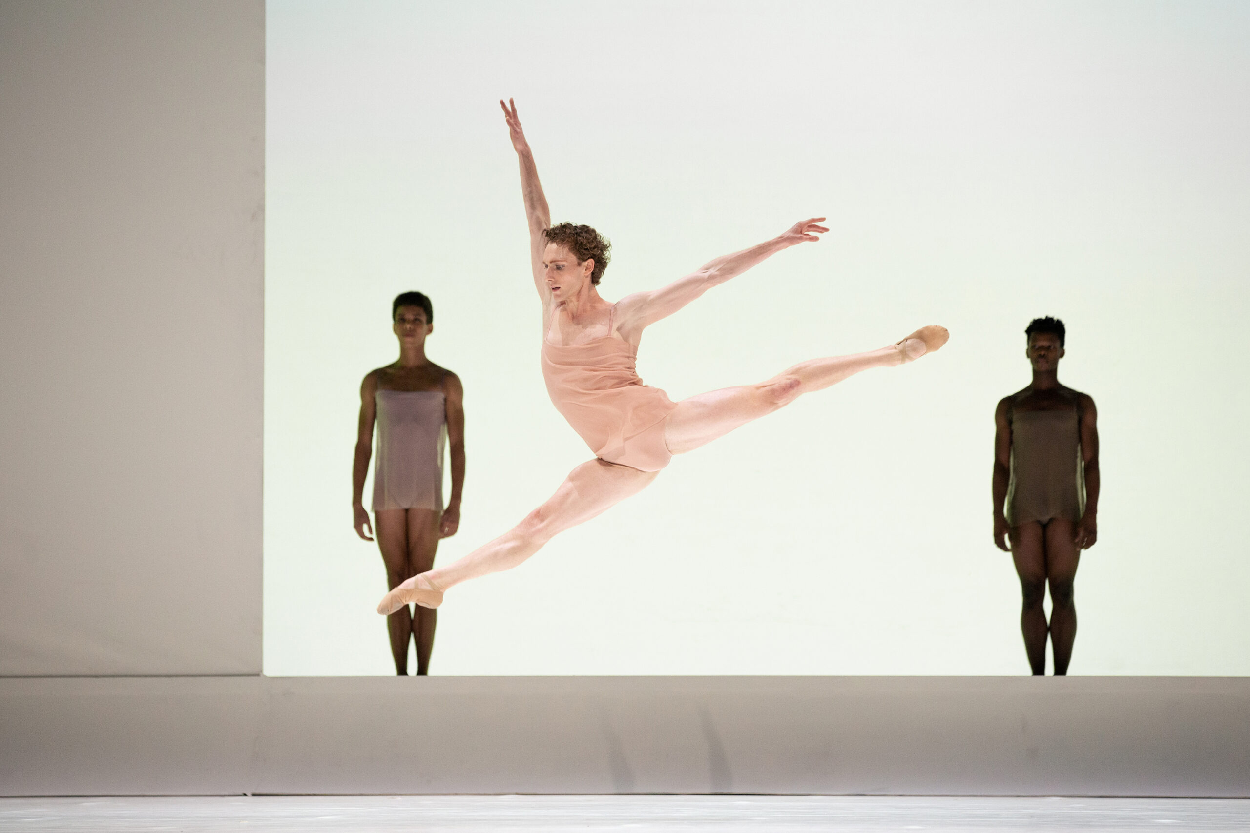 In front of a minimalistic set piece, Skylar Campbell splits his legs in the air with his arms in high arabesque. He wears briefs and a flowing top that match his skin tone. Two other dancers in similar costumes stand behind him on either side.