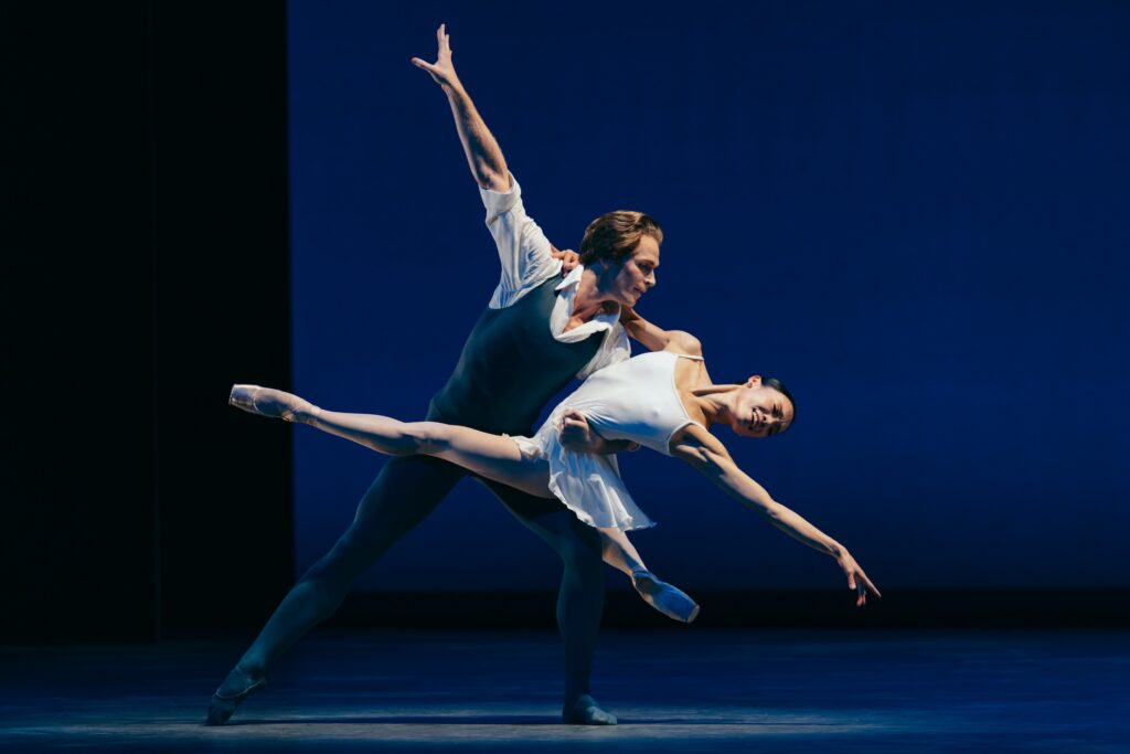 Two dancers perform a pas de deux in the spotlight of a somewhat darken stage. the man, wearing a white collared shirt under a dark blue unitard, lunges to his left side, his right arm extended high, as he holds the ballerina by the waist with his left arm. His partner, wearing a short white dance dress, wraps her right arm around his left shoulders and stretches her left arm long, her legs extended in a split over her partner's's left thigh. Both dancers look towards her left hand and smile.
