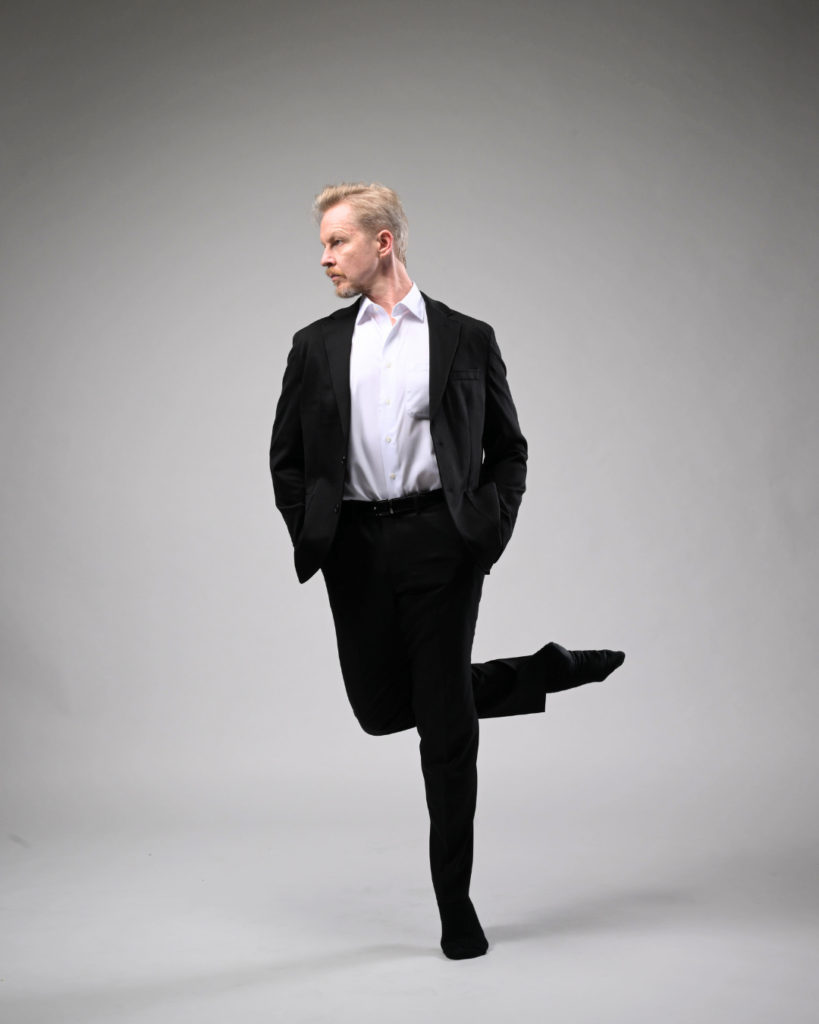 In front of a light gray backdrop, Tiit Helimets poses for a portrait in a formal black suit with a white button-down underneath. With his hands in his pockets, he looks to the left and tucks one leg up into an over-crossed passé, leaning left slightly.