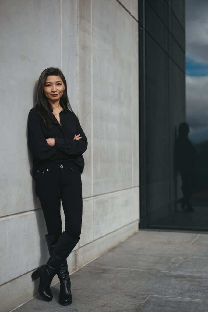 Tracy Li, wearing a dark blouse and jeans, leans back against a white stone wall with her arms crossed.