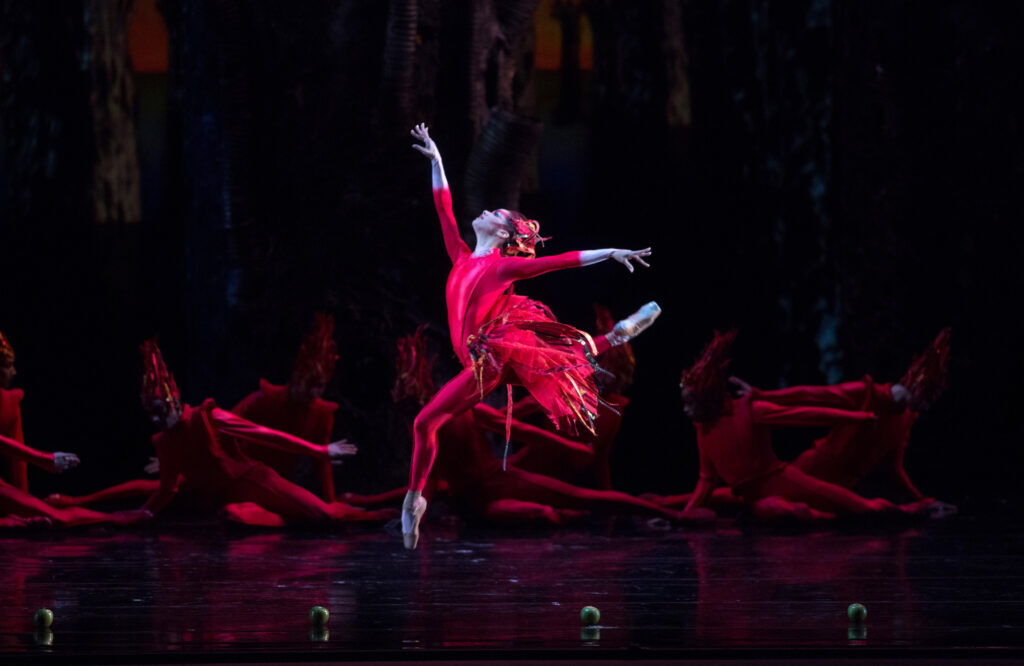 Misty Copeland leaps in profile, front and back legs in attitude, one arm reaching high overhead. She is costumed in a red unitard with a skirt and headress that evoke feathers, and pointe shoes pancaked to match her skin. Similarly costumed dancers recline on the floor upstage.