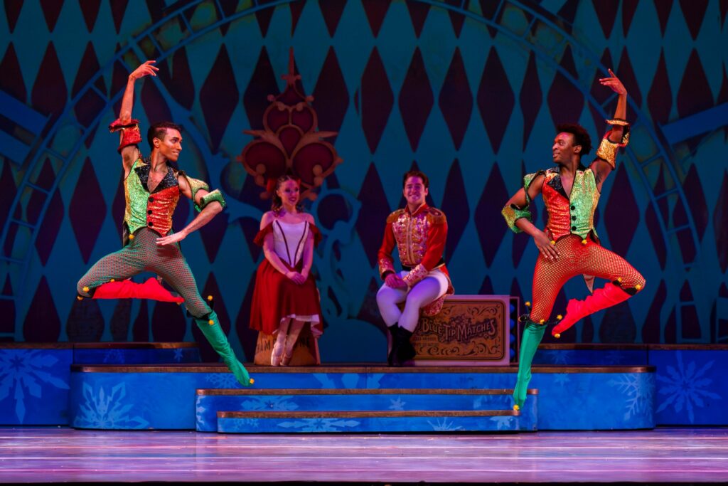 Two male dancers in red and green jester costumes dance opposite each onstage. They jump up with their outside leg in retiré and outside arm up, and their inside hand on their waist as they looks at each other, smiling. A young male and female dancer sit and watch in the background on a raised platform.