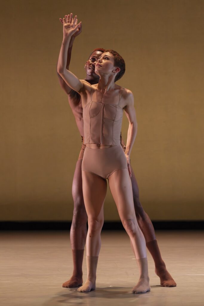 Lanie Jackson, wearing a taupe corset top and matching trunks and socks, stands onstage and lifts her right hand up, her left hand on her left hip. Her partner, Jared Kelly, wears brown shorts and socks and stands behind her, matching her movements.