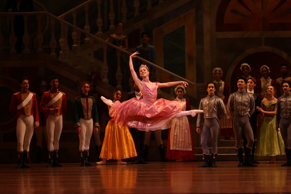 Tyler aDonatelli, wearing a long pink dress, tiara and pointe shoes, does a grand jeté with her back leg in attitude during a performance onstage. Her arms are in high third, and she looks out towards the audience with a big smile. A crowd of dancers in various artistocratic costumes stands in the background, watching.