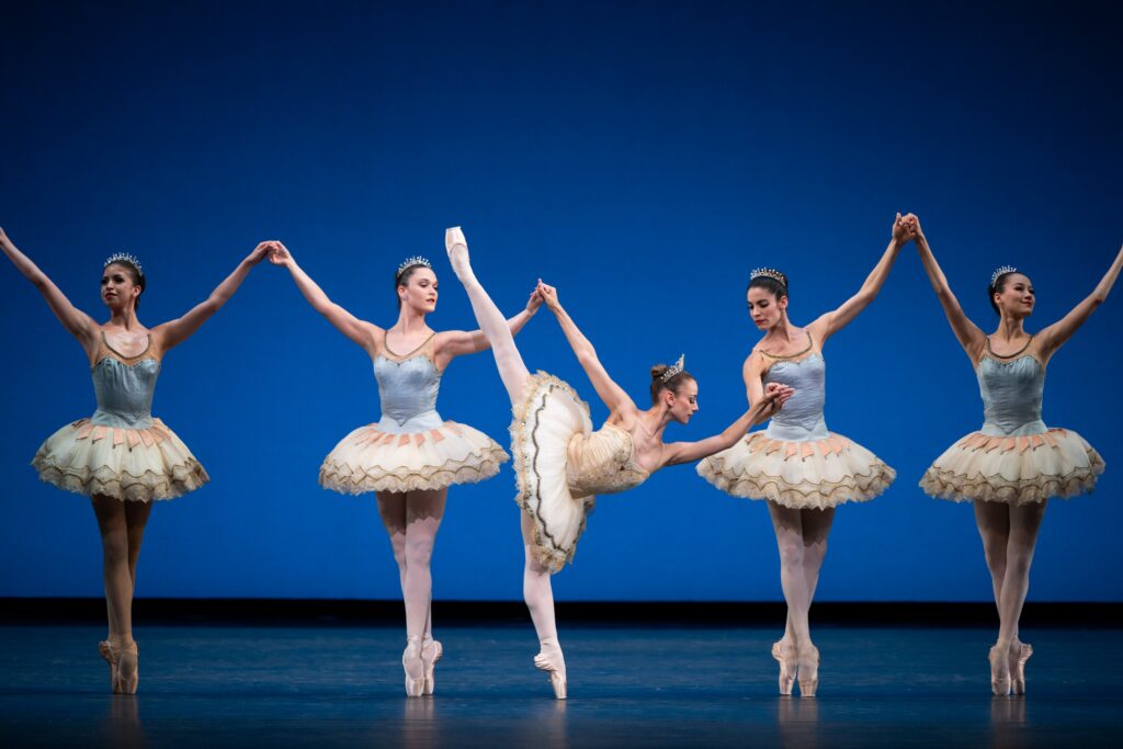 Tyler Donatelli does a penché on pointe while holding the hands of two dancers. The women are in a line of the corps, balancing in sus-sous. Donatelli wears a white and gold tutu and tiara, while the corps wear white tutus with blue bodices.