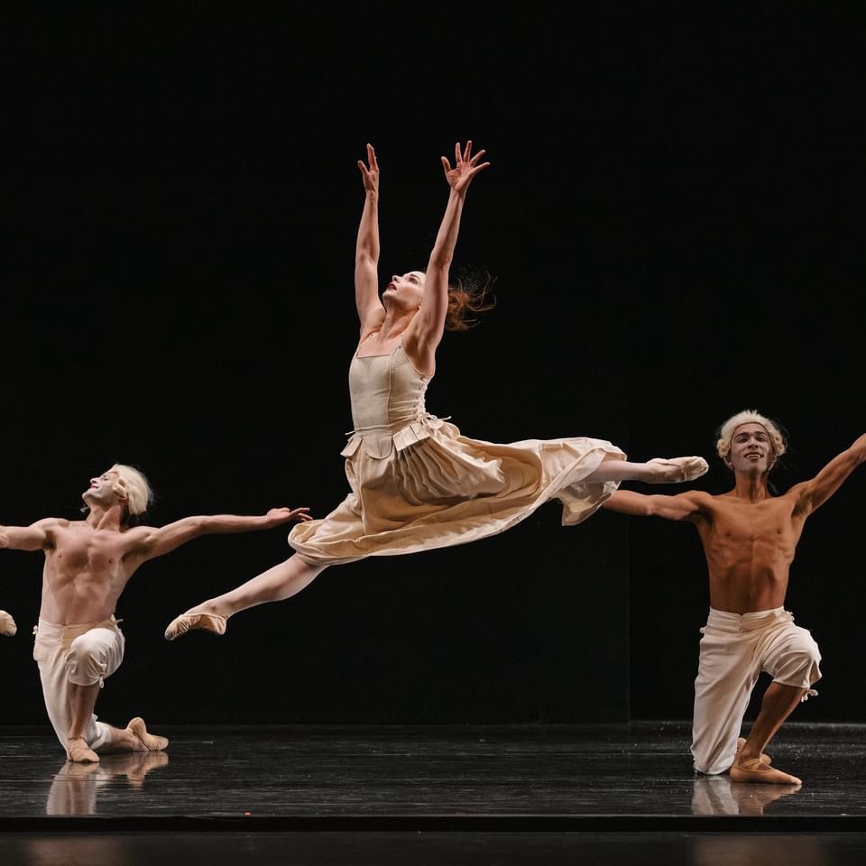 Chantielle Pianetta is captured mid-air, as she leaps onstage, her arms reaching straight-up above her head. She wears a beige dress and ballet shoes. Behind her, on either side, two male dancers pose on one knee, wearing light pants and colonial-style wigs.