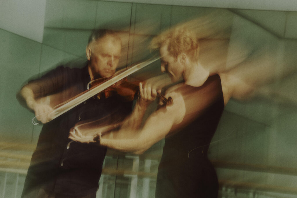 In a green tinted dance studio, Steven McRae and Vasko Vassilev are captured in motion from the waist up, their movements blurred. Vassilev, in a black button down and pants, is playing the violin facing forward. McRae, in a black tank top and pants, has his back to the camera as he faces Vassilev and gestures toward him with one arm.