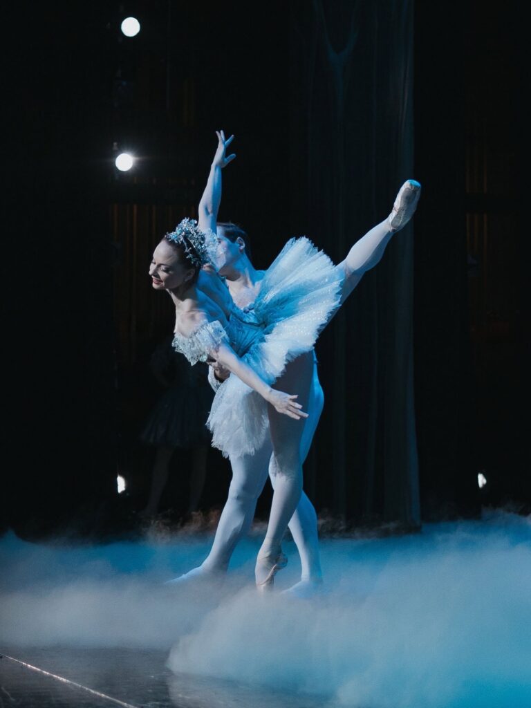 Chantelle Pianetta is captured onstage performing the snow pas de duex with Preston Swovelin. Pianetta is in an attitude penché en pointe, supported by Swovelin at her waist. Her arms reach out to her sides, palms turned away from her, and she looks down and over her left shoulder, which is nearest to the camera. She wears a white tutu and tall, glittery headpiece. The stage is filled with fog at the dancers' feet.