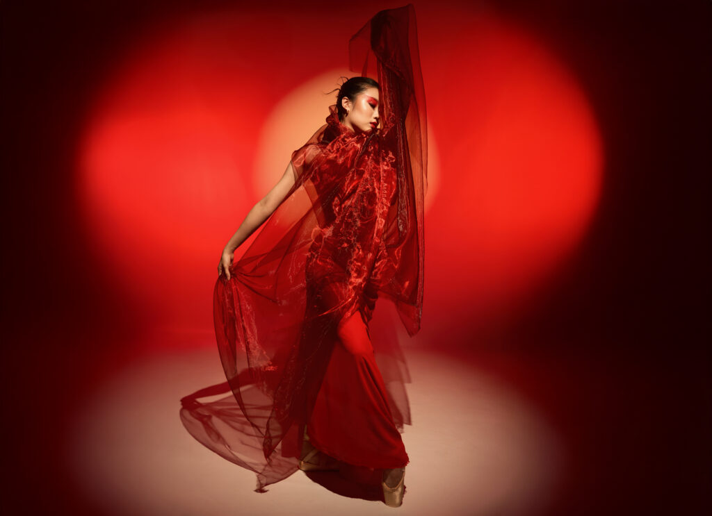 Sumin Lee is shown in a spotlight against a dramatic red background. She wears pointe shoes and a red costume of shiny draped fabic. She holds one of her arms, hidden by the fabric, above her head. Her other arm is by her side, gently holding the costume's overlay slightly away from her body. Lee has one foot crossed in front of her in a forced-arch and turns her head in profile, gazing downward, toward her lifted arm. She wears red eye-makeup that matches the backdrop and her costume.