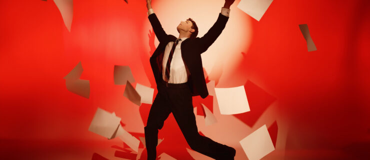 Logan Savard is shown against a red background with a spotlight haloing his upper body and head. He is wearing a suit: his jacket and pants are black, his dress shirt is white, and his tie has diagonal black-and-red stripes. Savard is looking upward with his arms thrown in the air above his head, palms facing out. He is surrounded by a flurry of blank pieces of paper.