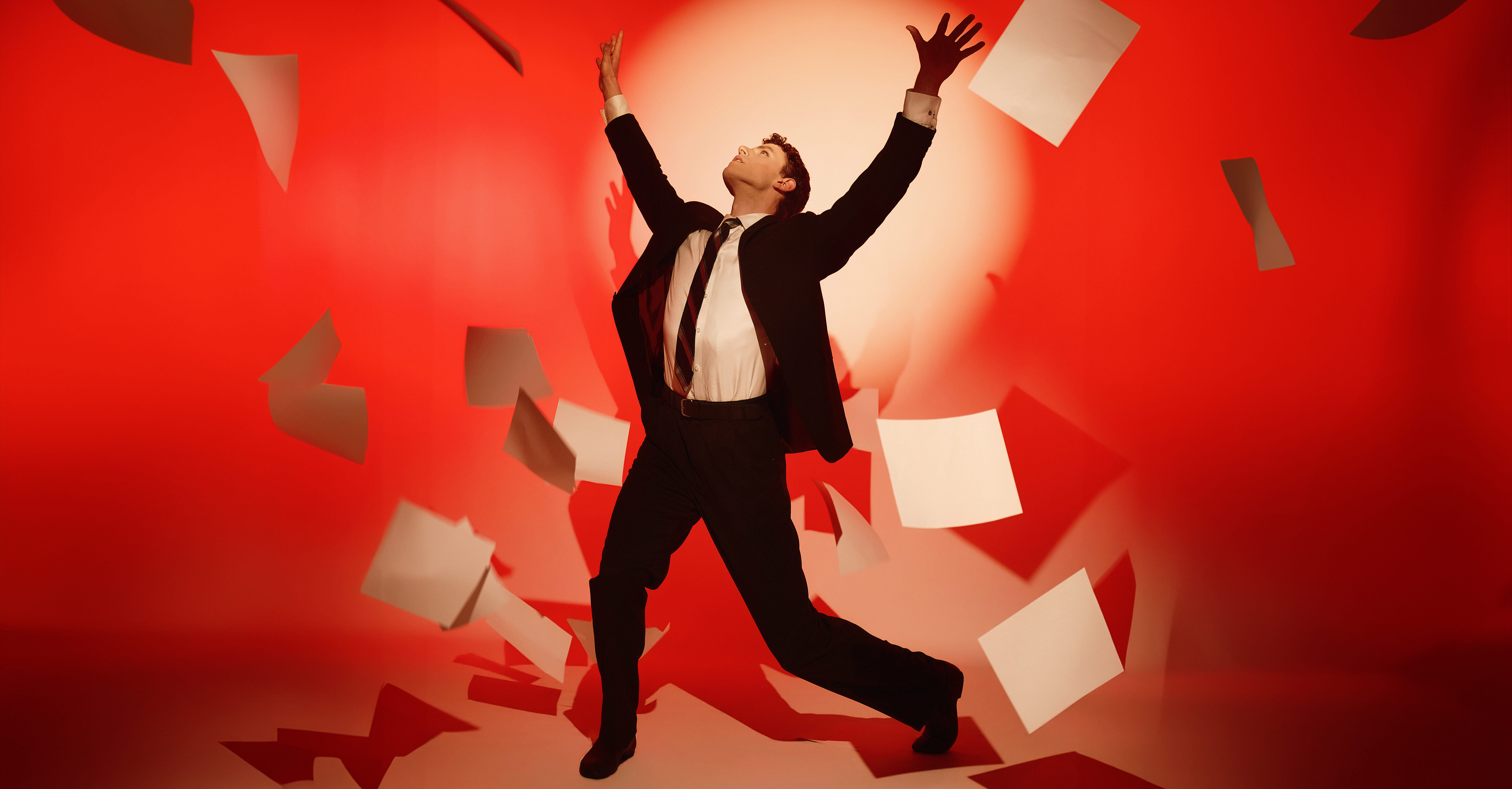 Logan Savard is shown against a red background with a spotlight haloing his upper body and head. He is wearing a suit: his jacket and pants are black, his dress shirt is white, and his tie has diagonal black-and-red stripes. Savard is looking upward with his arms thrown in the air above his head, palms facing out. He is surrounded by a flurry of blank pieces of paper.