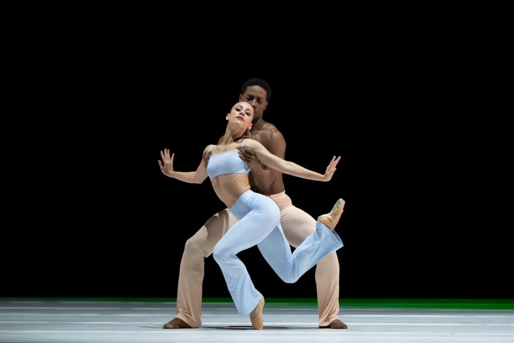 Eric Best and Tyler Donatelli perform a contemporary pas de deux onstage in front of a black backdrop. Donatelli wears a light blue bandeau top and matching pants, and poses on pointe in a low crouch on her left foot, her right leg lifted and her arsm open, wrists flexed. Best, wearing tan pants, stands behind her in second position and holds her under the armpits.