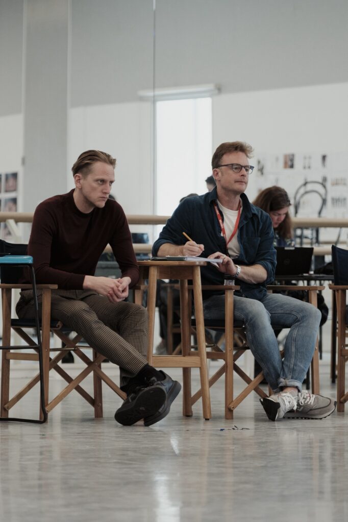 David hallberg and Christopher Wheeldon sit in chairs at the front of a dance studio and observe a rehearsal. They both ear streest clothing, and Wheeldon takes notes.