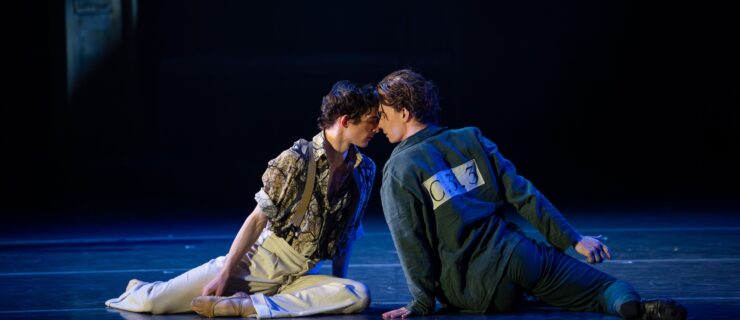 Dancers Jarryd Madden and Adam Elmes sit on the stage floor during a performance, facing each other and touching foreheads.