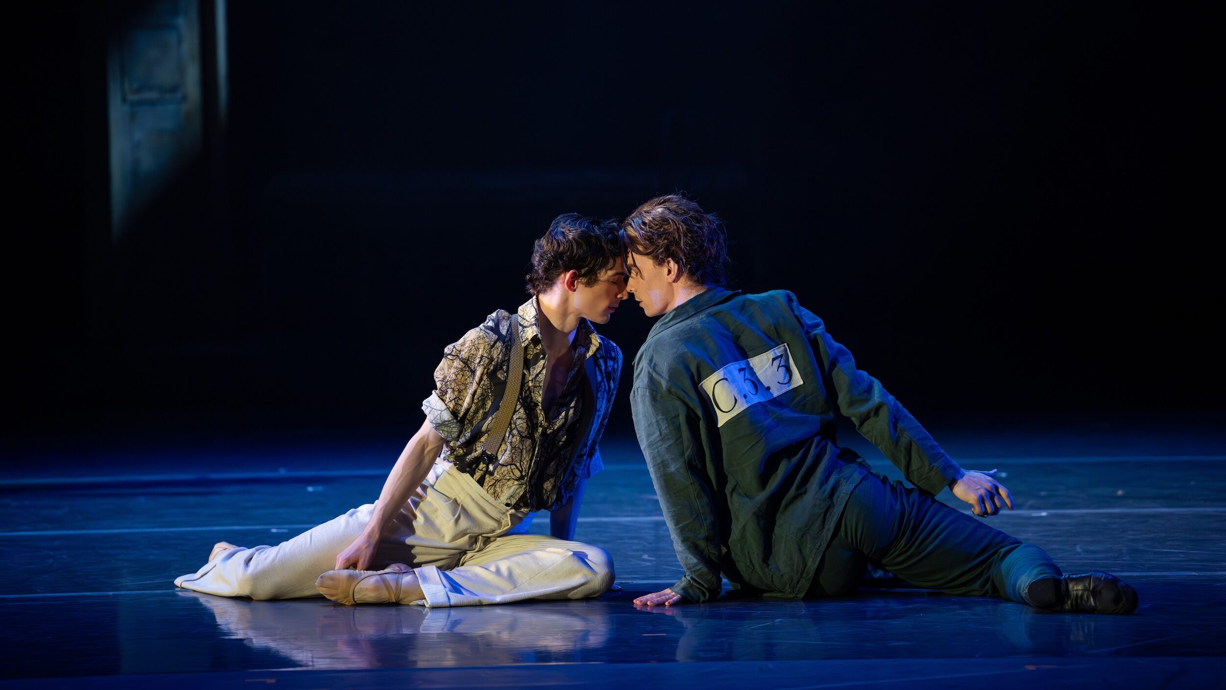 Dancers Jarryd Madden and Adam Elmes sit on the stage floor during a performance, facing each other and touching foreheads.