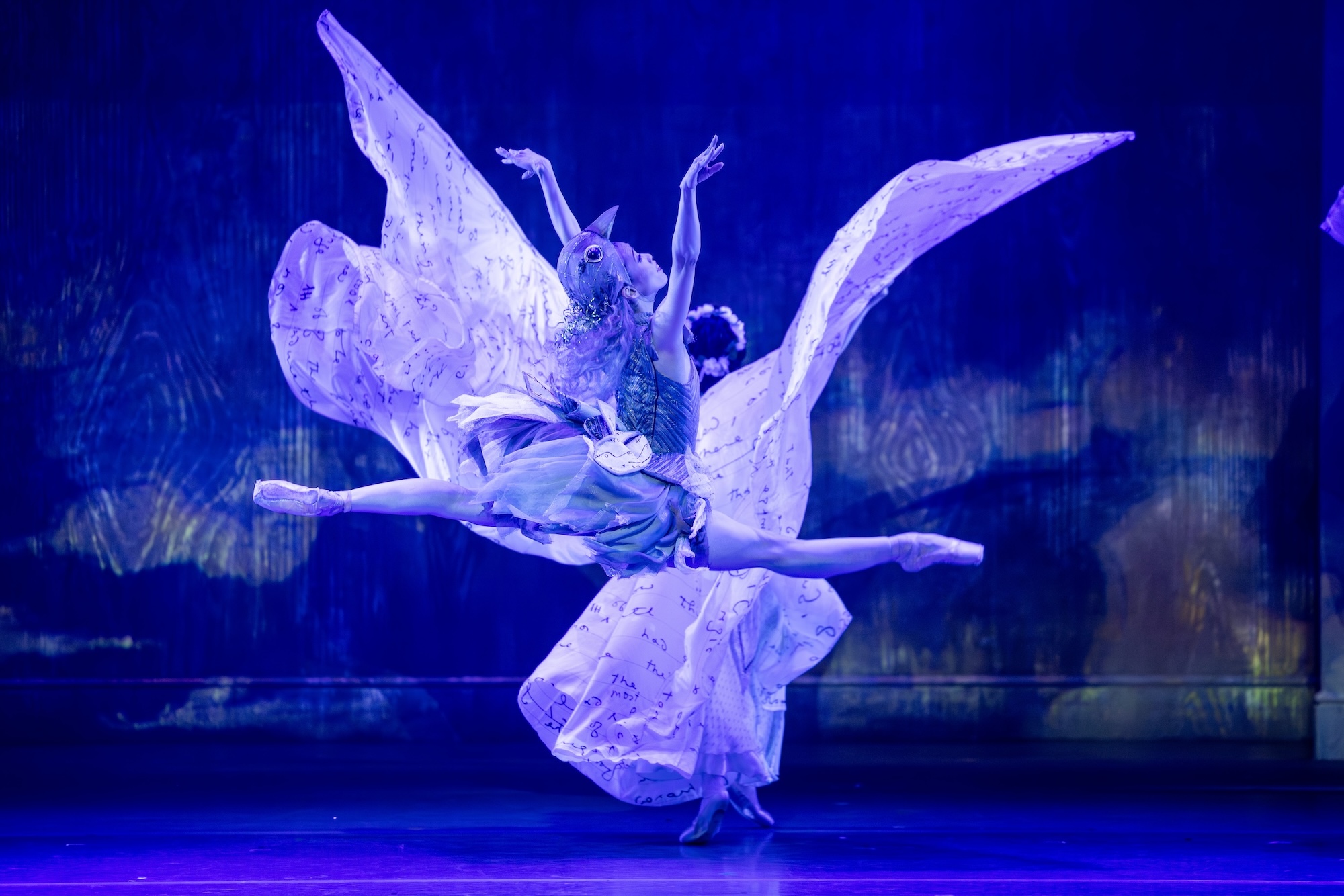 Ako Kondo, bathed in purple ight, performs a saut de chat upstage with her arms raised in a V shape. She wears a bird costume that consists of a knee-length dress and a headpiece. Another dancer upstage wears a long costume with giant sleeves and throws her arms in the air so that the sleeves look like wings.