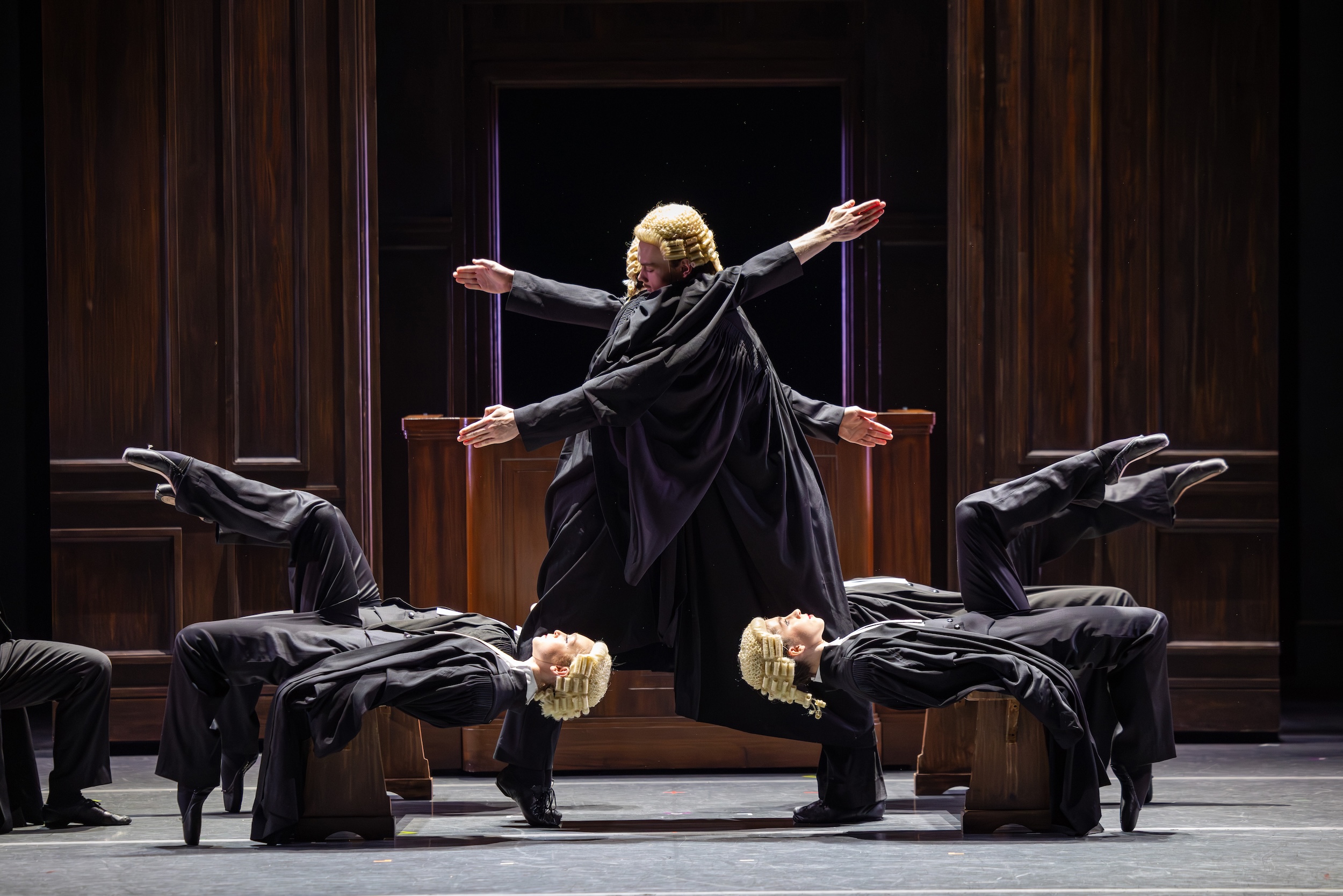 A corps of dancers dressed like English judges wear black suits, long cloacks, and powdered wigs. two leab into each other with their arms outstretched while the others lay on their backs on top of court benches with their legs raised in table top positions.