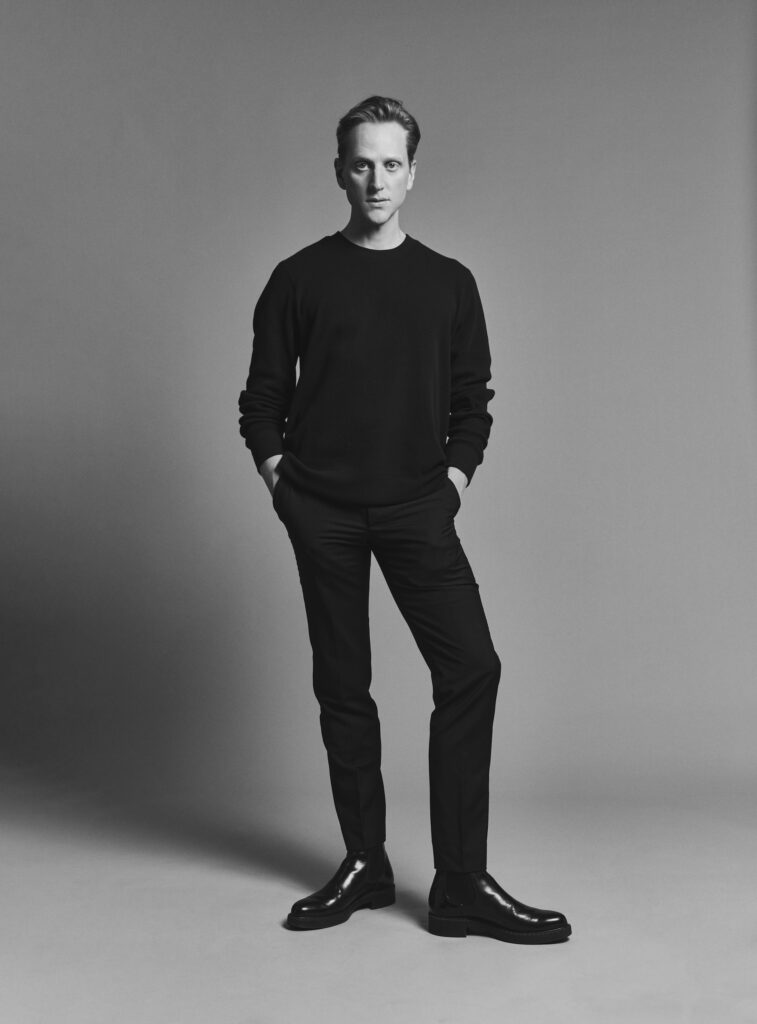 David Hallberg poses for a black and white portrait. He stands with his hands in his pockets and with his left foot slightly turned out. He wears a black sweater, pants, and dress shoes and looks directly toward the camera.
