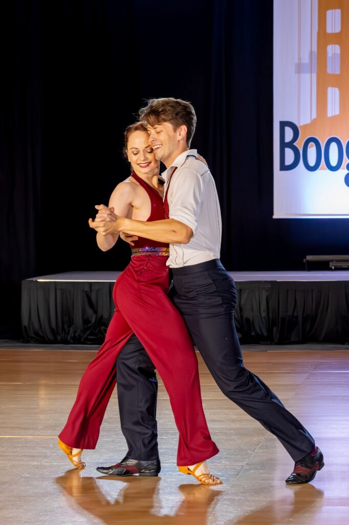 Chantelle Pianetta dances with Joel Torgeson on wooden flooring. Pianetta wears a red jumpsuit with tan ballroom shoes. Torgeson wears a white dress shirt with black pants and dress shoes. Both dancers smile, their heads pressed close together and turned toward their clasped hand.