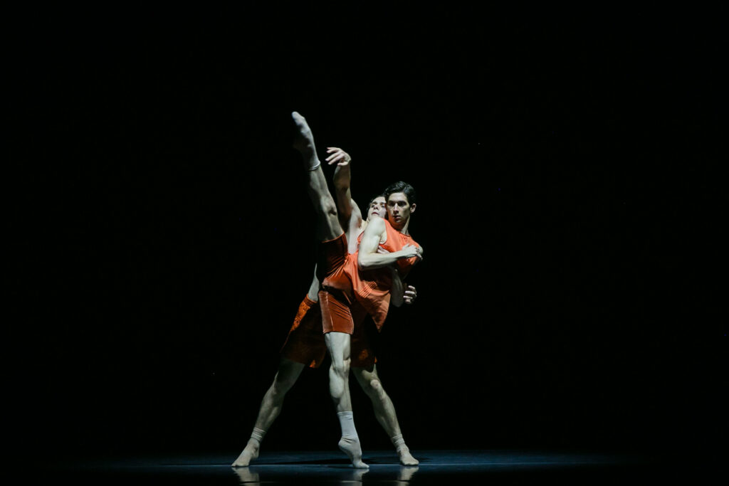Adam Elmes and Callum Linnane are shown dancing together onstage against a black backdrop, Elmes in front of Linnane. Elmes is on relevé with one leg extended to the side, above their heads. Linnane supports him from behind, with one arm reaching toward the lifted leg. Both dancers wear orange shorts and light-colored socks. Elmes also wears a matching orange tank top.