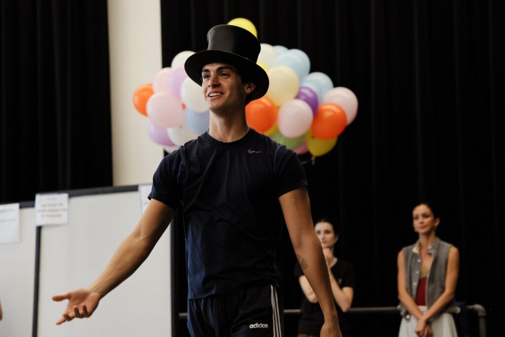 José Pablo Castro Cuevas is shown from the waist up wearing a dark t-shirt and top hat. He extends his right hand out, palm up and smiles. In the background, two dancers stand behind him, and an assortment of colorful balloons frame his head.